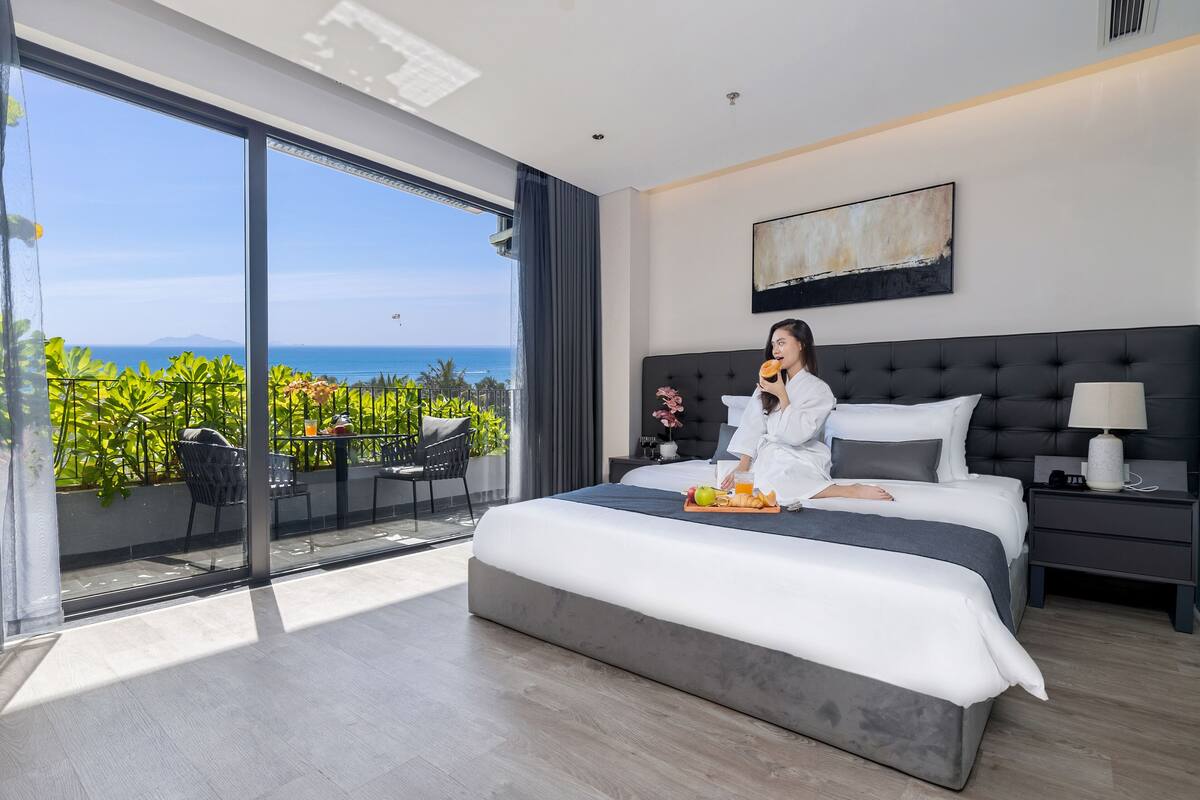 Une chambre d’hôtel moderne dotée d’un grand lit, d’une table de chevet, d’une commode et offrant une vue sur l’océan grâce à de grandes fenêtres.