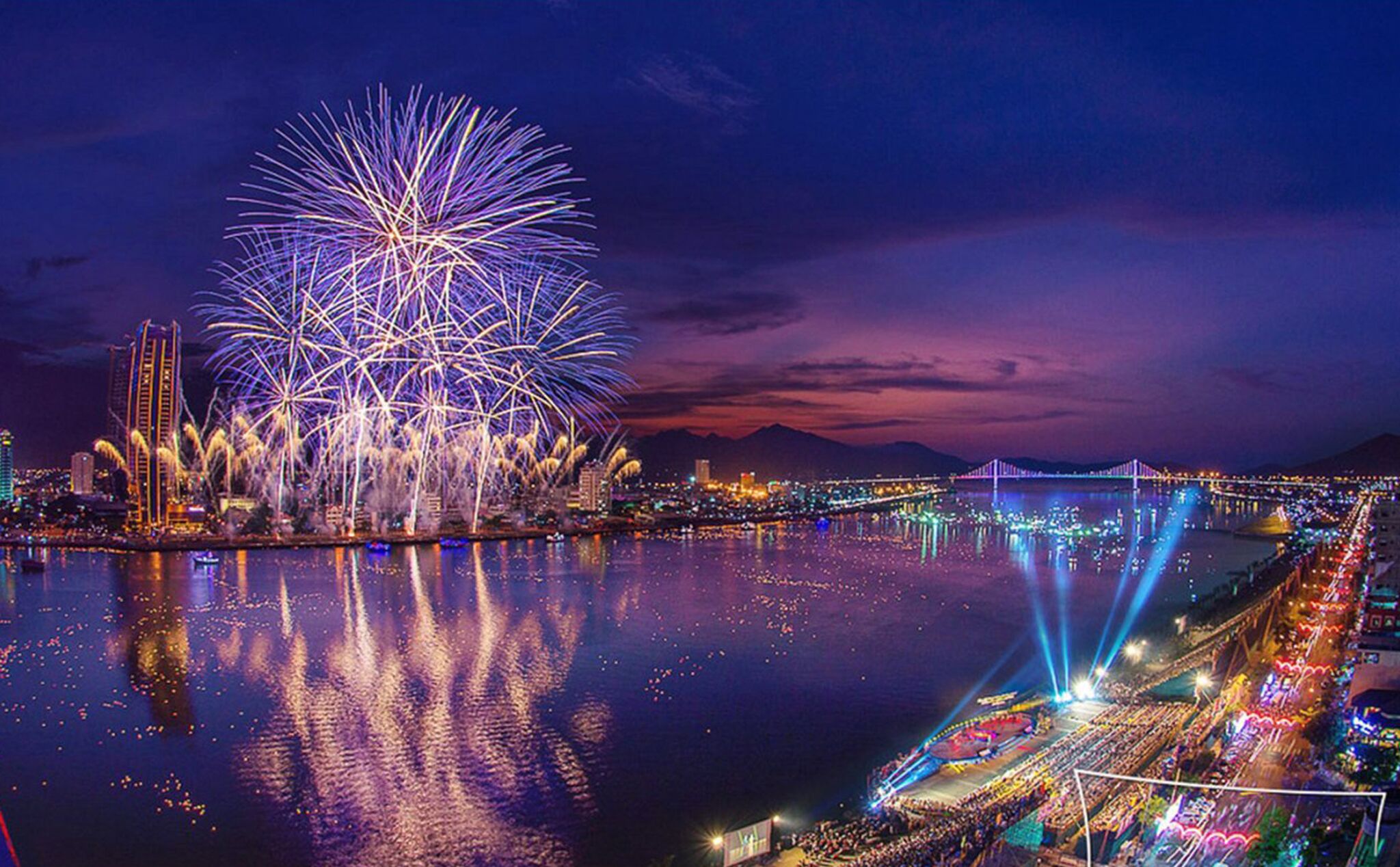A waterfront cityscape with fireworks over a body of water, a bridge, and a stadium.