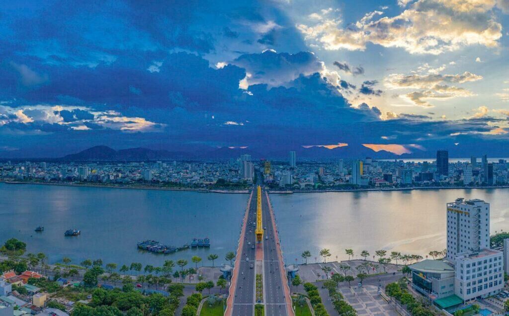 A cityscape with a prominent bridge leading to a body of water, surrounded by buildings and greenery.