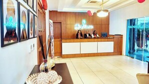 A hotel reception area with a wooden reception desk, framed artwork on the walls, and a large window offering a view of the outdoors. - De Palms Hotel Port Harcourt (Port Harcourt)