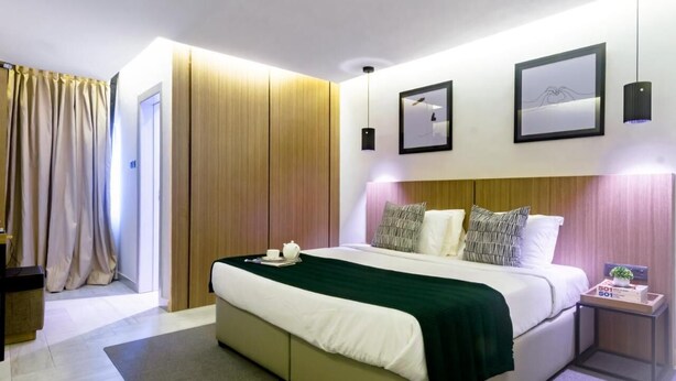 A modern hotel room with a large bed, a bedside table with a plant, and a view of the city. - De Palms Hotel Port Harcourt (Port Harcourt)