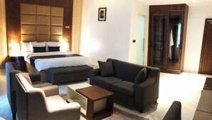 A hotel room with a large bed, two sofas, a coffee table, and a small armchair. There is a wooden headboard, a framed picture on the wall, and a door with a mirror. - De Palms Hotel Port Harcourt (Port Harcourt)