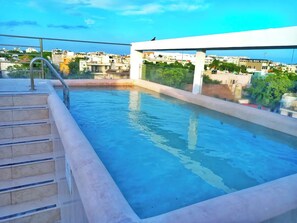Ein Pool auf dem Dach mit Stufen zum Wasser, umgeben von einer weißen Struktur und Blick auf die Stadtlandschaft.