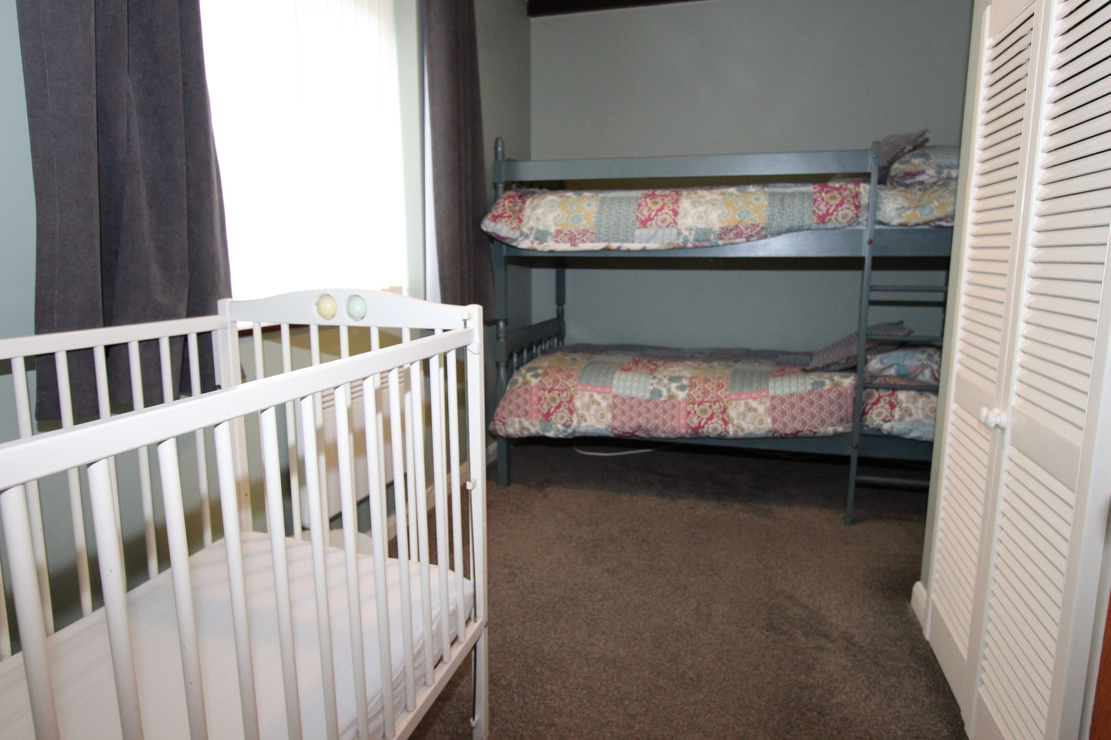 A room with a white crib, a bunk bed with floral bedding, and a closet with louvered doors.
