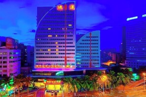 A multi-story hotel with a curved facade, illuminated signage, and a parking area with palm trees. - Hainan Hongyun Hotel (Haikou Qilou Old Street) (Haikou)