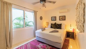 Une chambre à coucher comprenant un lit, un ventilateur de plafond, une fenêtre avec des rideaux, une table de chevet avec une lampe et des éléments de décoration murale.