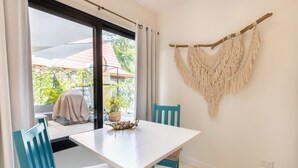 Habitación con una mesa blanca, sillas azules, un colgador de macramé y vista a un área exterior cubierta con plantas.