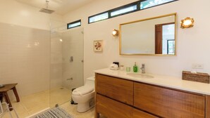 Un baño con cabina de ducha de vidrio, inodoro, tocador con espejo y un taburete pequeño.