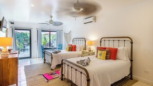 Habitación de hotel con dos camas, ventilador de techo, unidad de aire acondicionado, escritorio con lámpara y vista al exterior a través de una puerta de cristal.