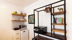 Cocina compacta con microondas, un refrigerador pequeño y un basurero. Hay un estante de madera con un cuadro enmarcado y una caja fuerte negra.