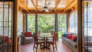 A covered patio with a wooden ceiling, a ceiling fan, a round table with chairs, and a sofa.