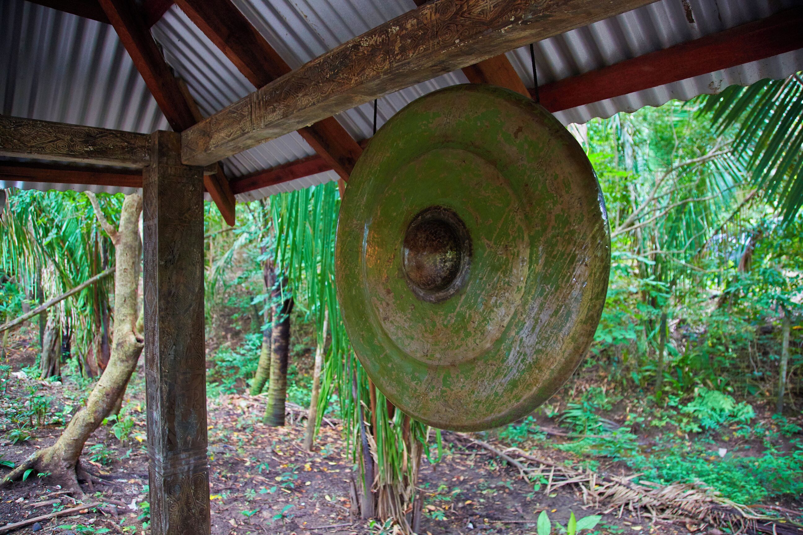 A large green gong hanging from a wooden structure in a forested area.