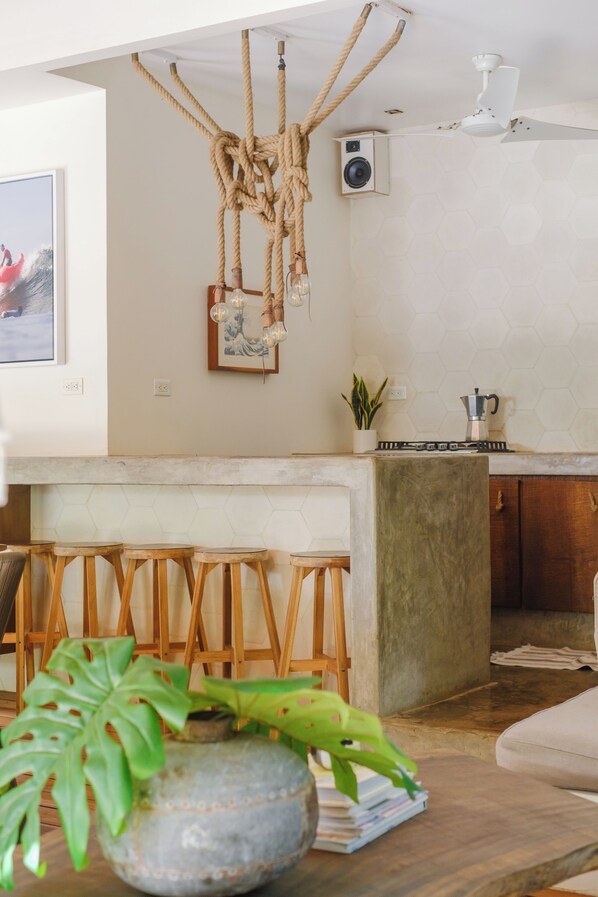 A modern kitchen with a wooden bar stool seating area, a hanging rope light fixture, and a potted plant on the counter.