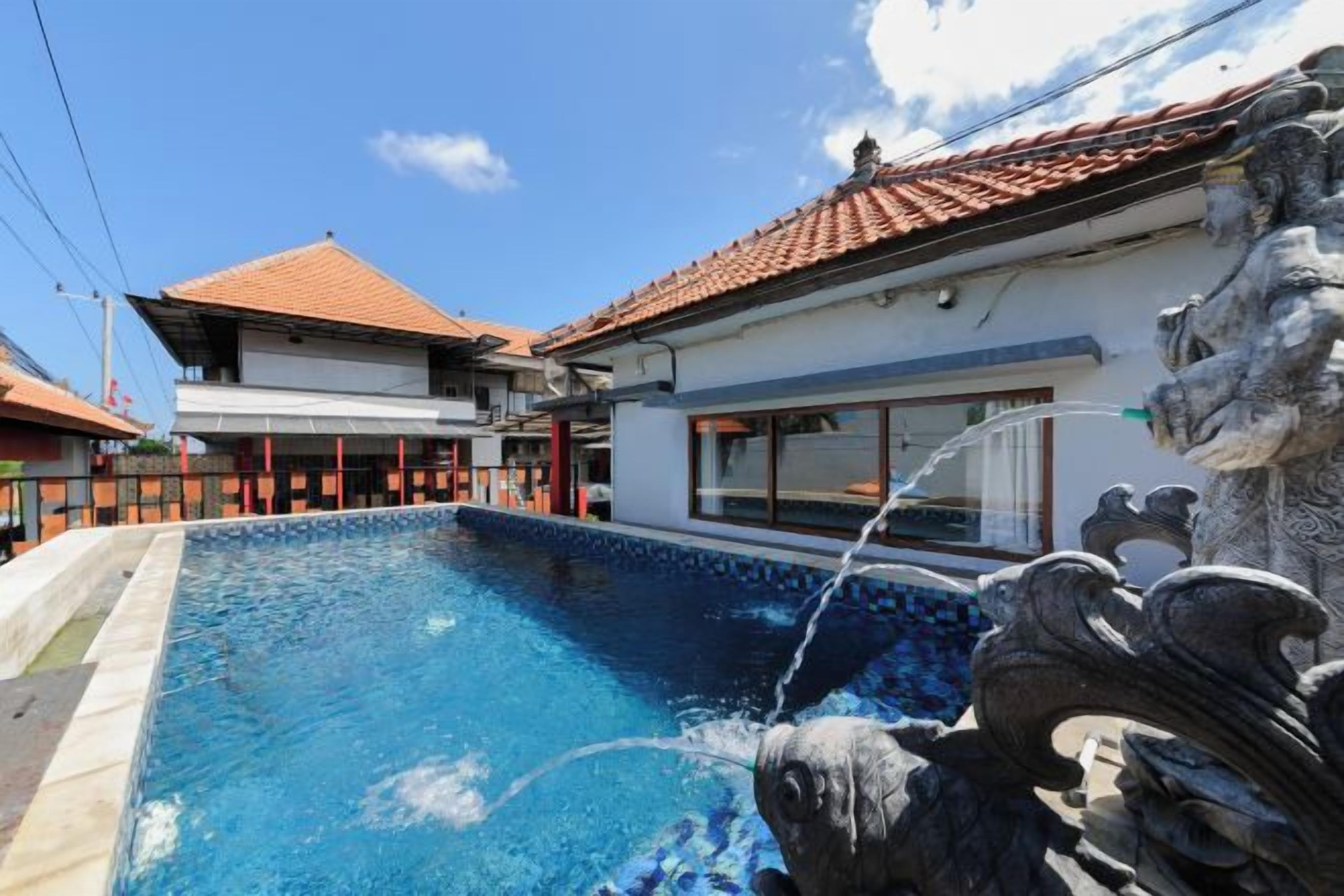 Ein Schwimmbecken mit Wasserspiel, umgeben von einem Gebäude mit einem Ziegeldach und einer Steinstatue.