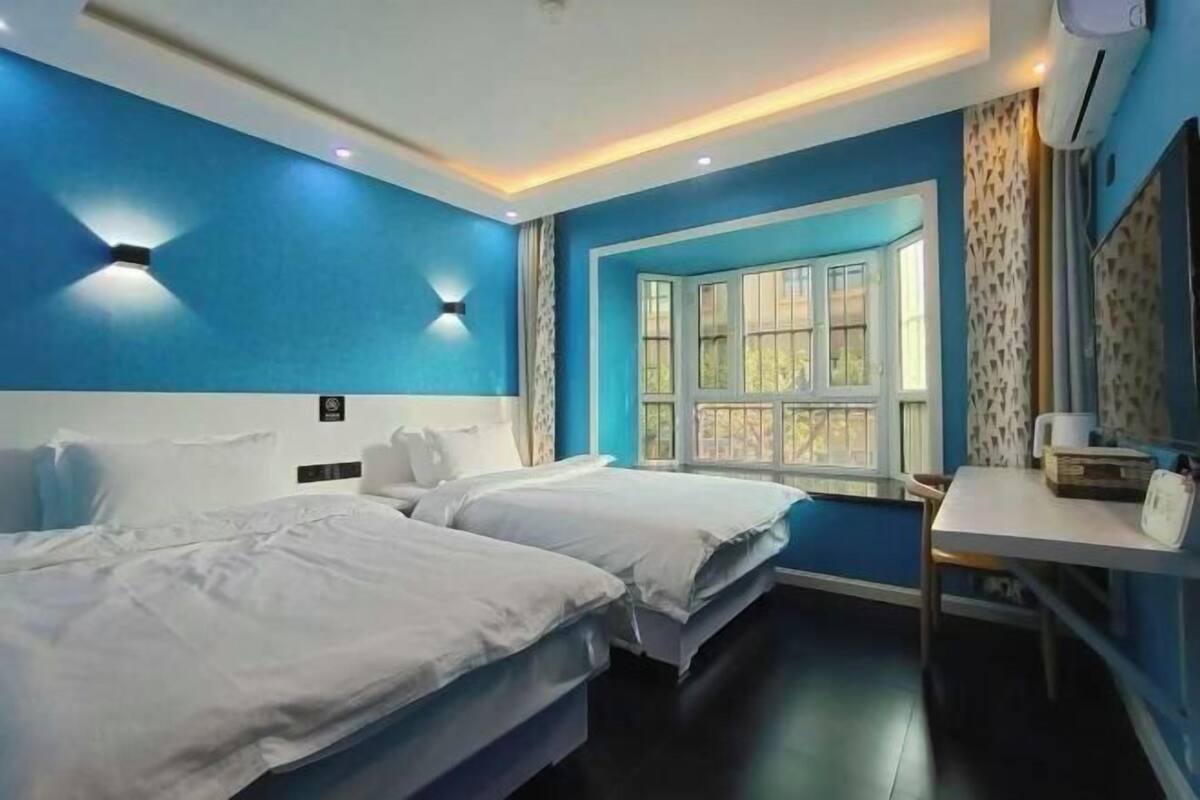 A hotel room with two beds, a small table, a window with curtains, and a wall-mounted air conditioner.