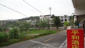 A red sign with Chinese characters for a hotel, a road with a bridge in the background, and buildings on the hillside. - Yongshun Pinghe Homestay (Yongshun)