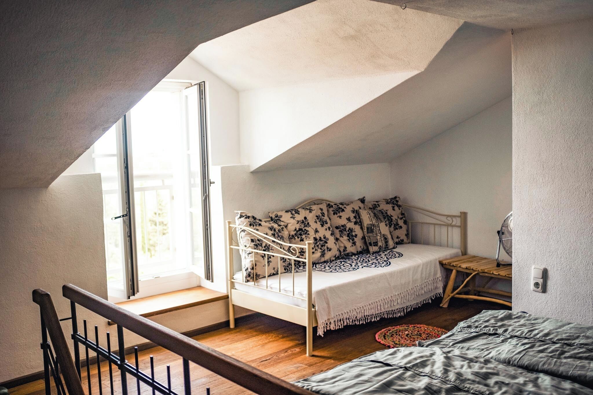 A bedroom with a sloped ceiling, a bed with a floral-patterned headboard, a small wooden table, and a window with a view of the outdoors.