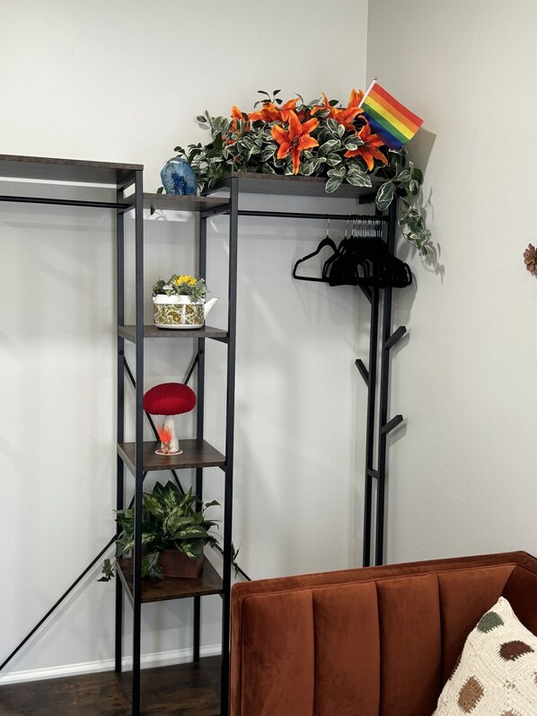 Un rincón con una estantería metálica negra que contiene diversos objetos decorativos, incluyendo una bandera arcoíris, una lámpara de seta roja y plantas en macetas.
