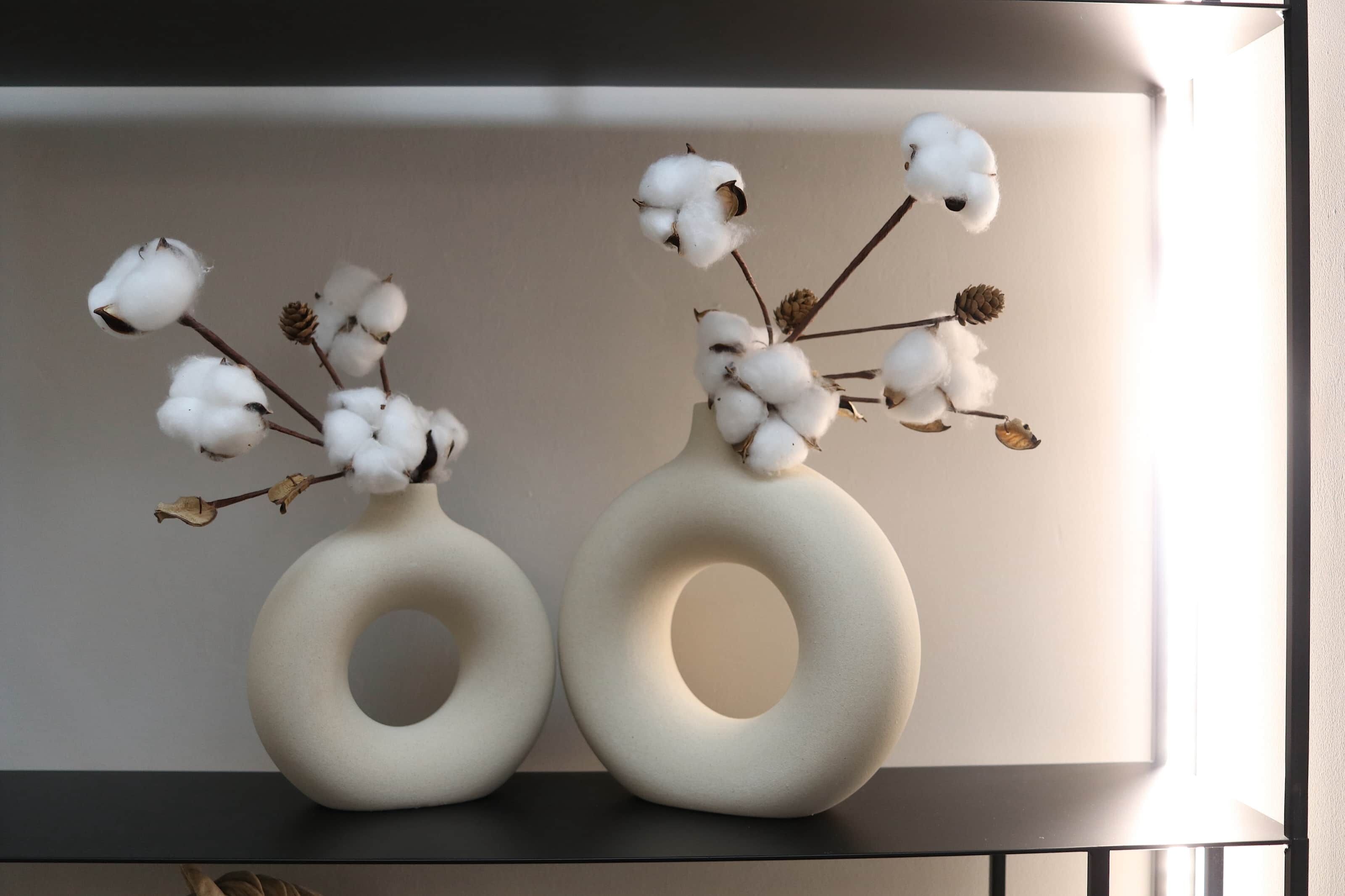 Two white, circular vases with cotton branches and dried leaves on a shelf.