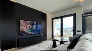 Una sala de estar moderna con un televisor de pantalla plana grande montado en una pared oscura, un sofá con cojines estampados, una mesita redonda y vistas al exterior a través de una puerta de cristal.
