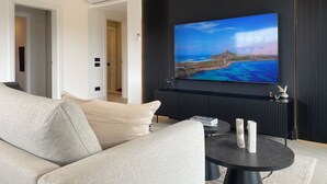Una sala moderna con un televisor de pantalla plana grande montado en una pared oscura, un sofá color beige y una pequeña mesa de centro redonda con objetos decorativos.