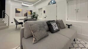 Ein modernes Wohnzimmer mit einem grauen Sofa, gemusterten Kissen, einem Essbereich mit Stühlen und einer Treppe.