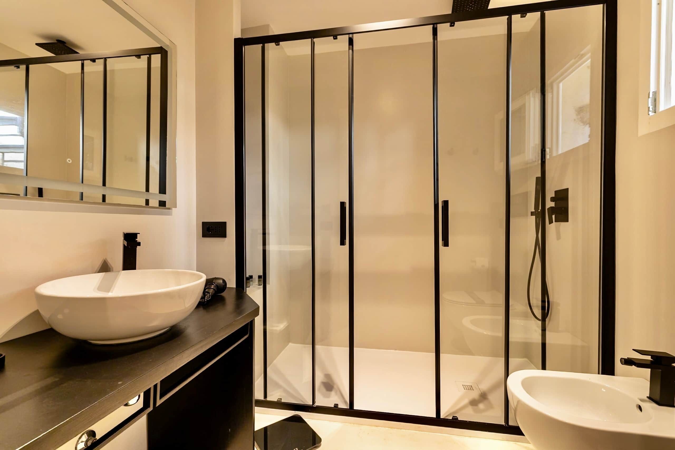 Un baño moderno con un espejo grande, un lavamanos tipo cuenco blanco, un lavamanos independiente y una zona de ducha con paredes de vidrio.