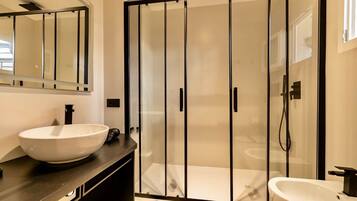 Un baño moderno con un espejo grande, un lavamanos tipo cuenco blanco, un lavamanos independiente y una zona de ducha con paredes de vidrio.