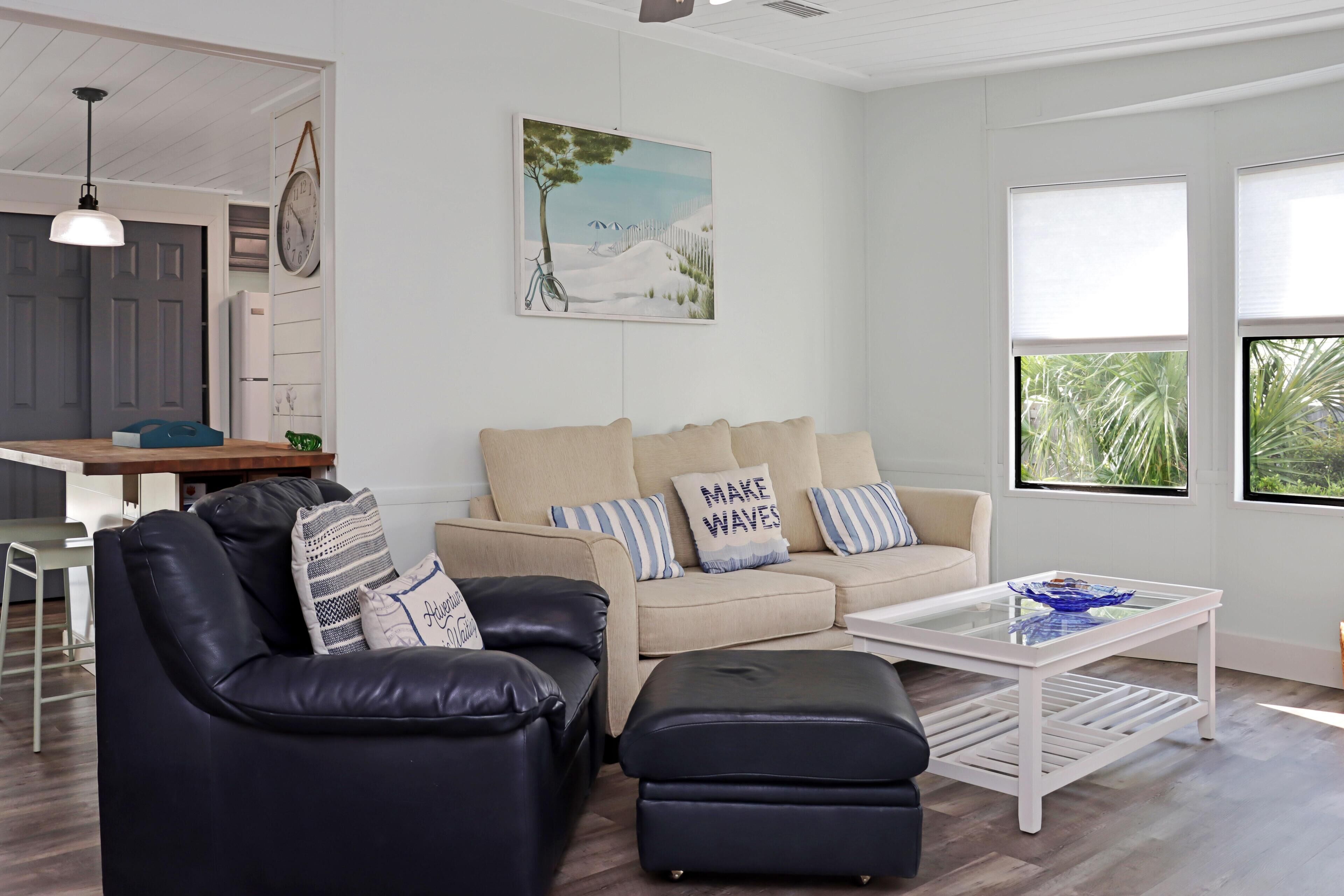A living room with a black leather sofa, a beige sofa, and a glass coffee table. There is a painting on the wall and a clock on the wall.