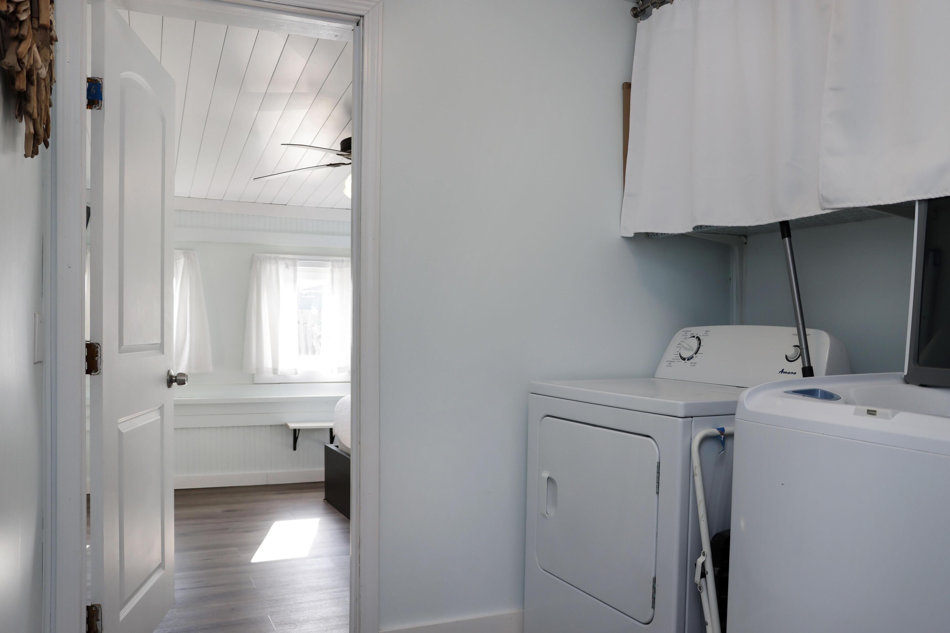 A laundry room with two white washing machines, a drying rack, and a door leading to a room with a wooden floor and a window with curtains.