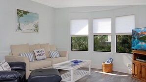 Een moderne woonkamer met een beige bank, zwarte leren poefjes, een glazen salontafel en een televisie op een houten dressoir.