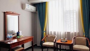 Habitación de hotel con un escritorio de madera, dos sillas, una mesita con tetera y un ventanal grande con cortinas amarillas y azules.