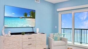 Een kamer met een grote tv waarop een strandafbeelding te zien is, een wit bergingsmeubel met lades, een lichtkleurige fauteuil met een gekleurd kussen, en een groot raam met balkon.