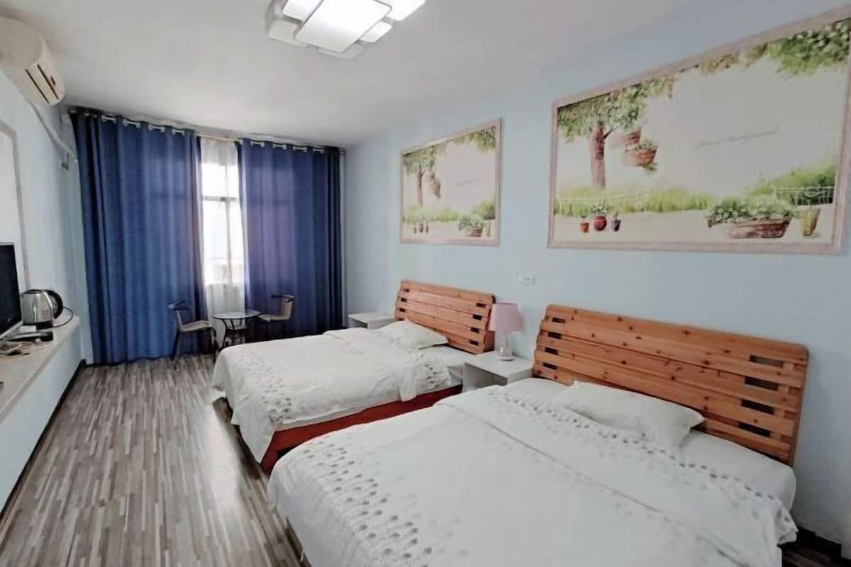 A hotel room with two beds, a desk with a chair, a small table, and a window with blue curtains.