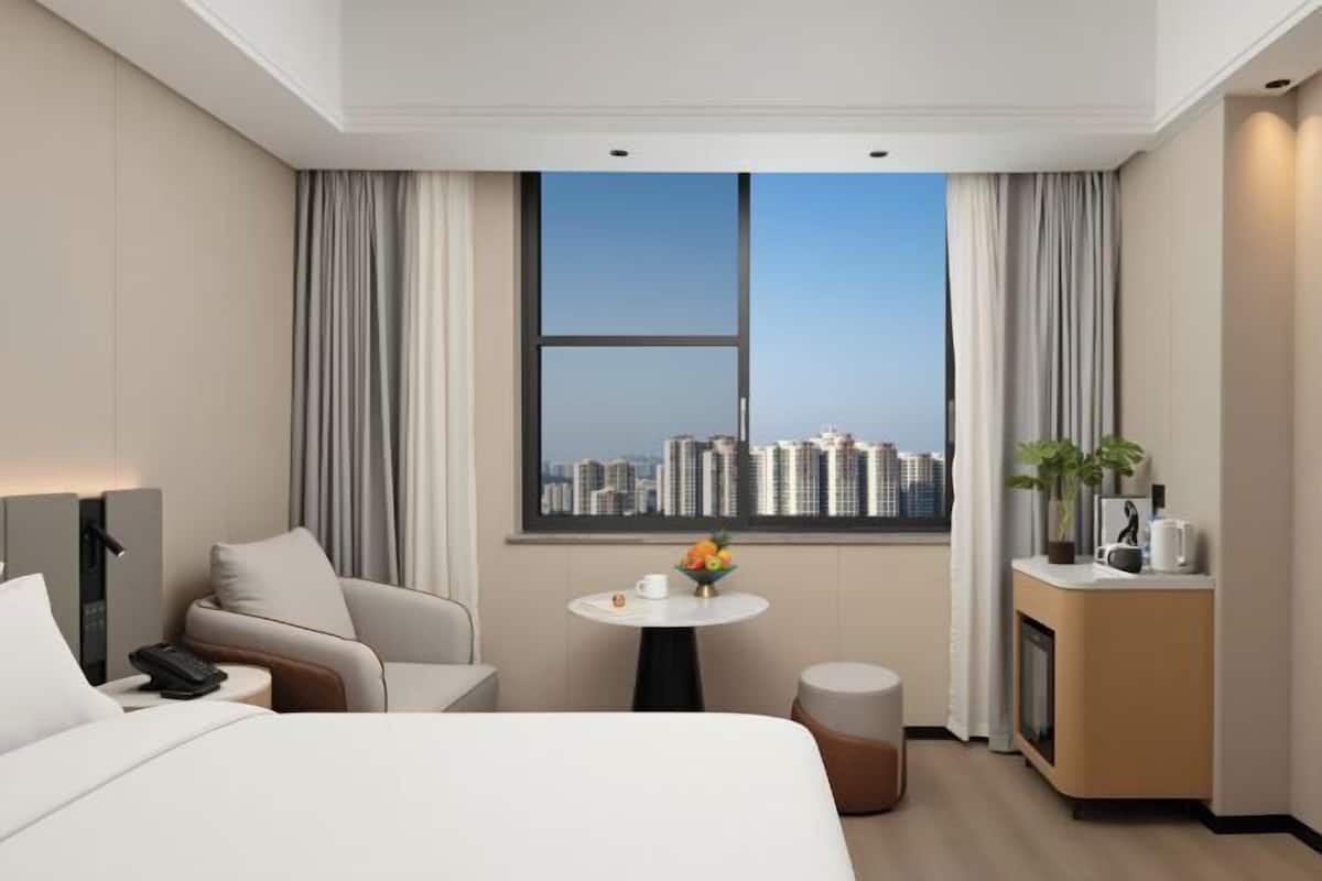 Quarto de hotel com cama, área de estar, mesa pequena com um vaso, escrivaninha com planta e janela com vista para a cidade.