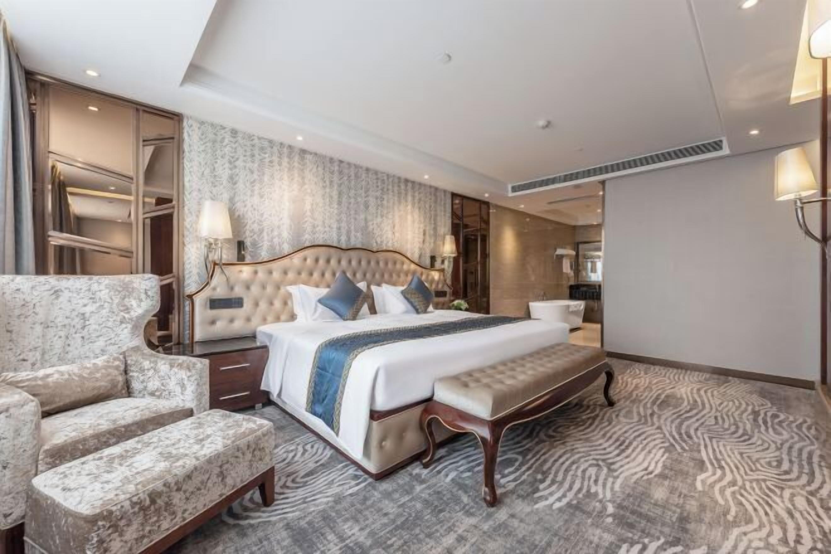 A hotel room with a large bed, a seating area with a bench, a wardrobe, and a mirror.