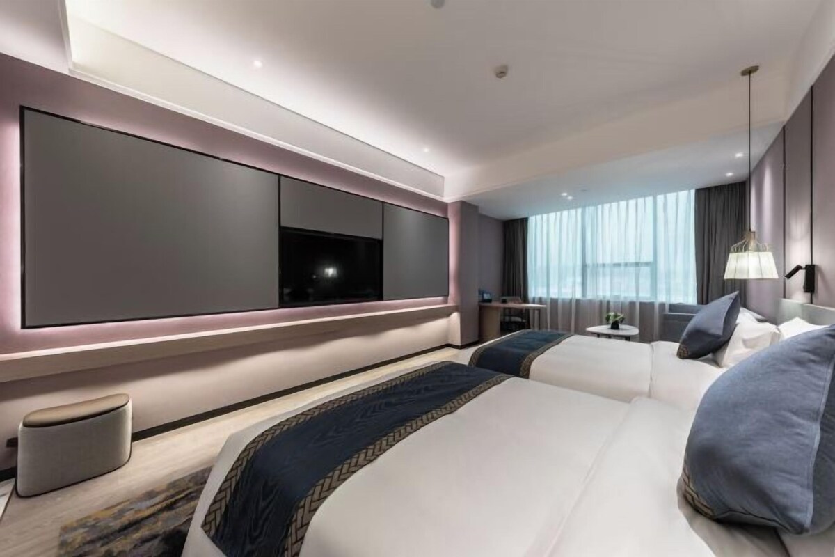 A modern hotel room with a large bed, a TV mounted on the wall, a desk, and a window with curtains.