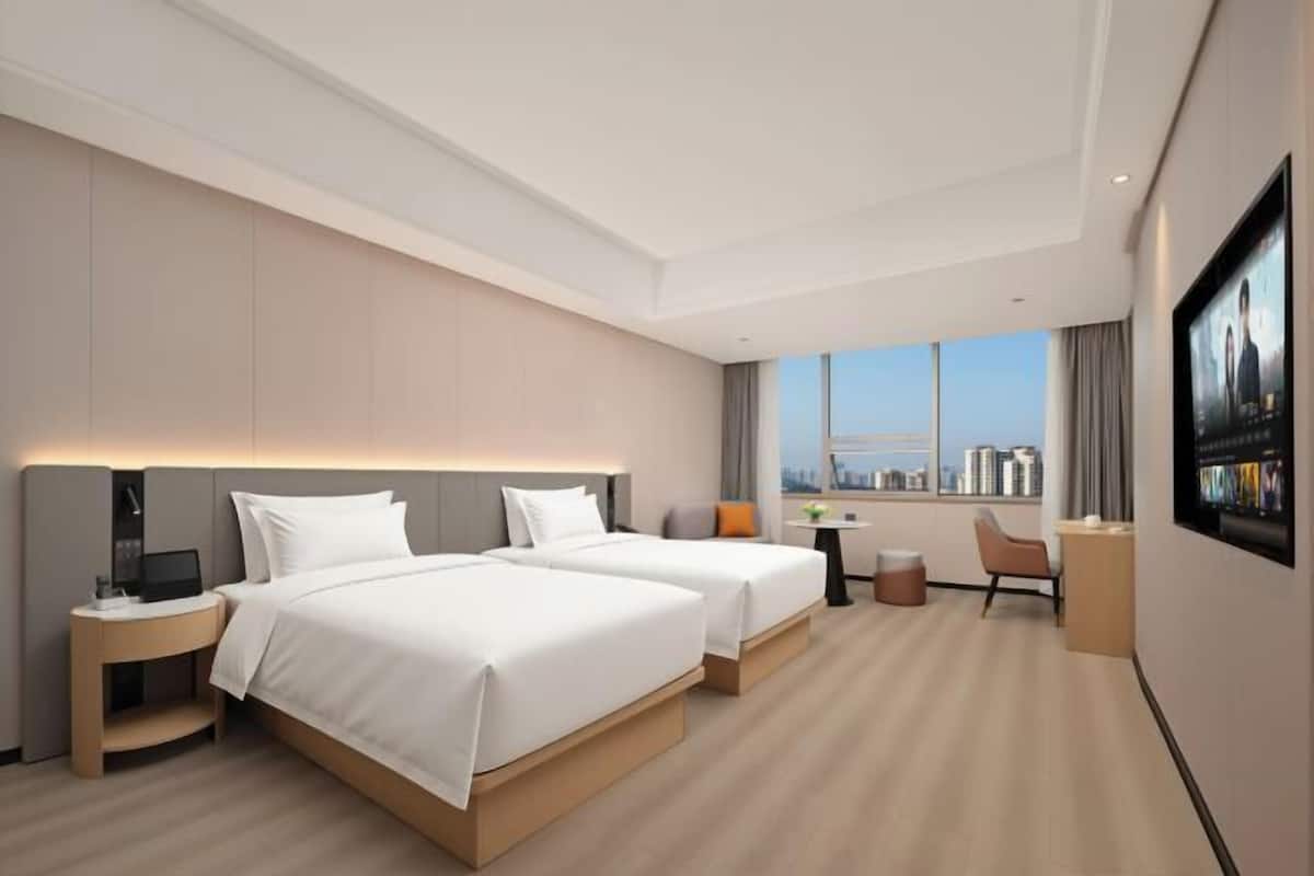 Quarto de hotel com duas camas, uma escrivaninha, uma cadeira, televisão e vista para a cidade.