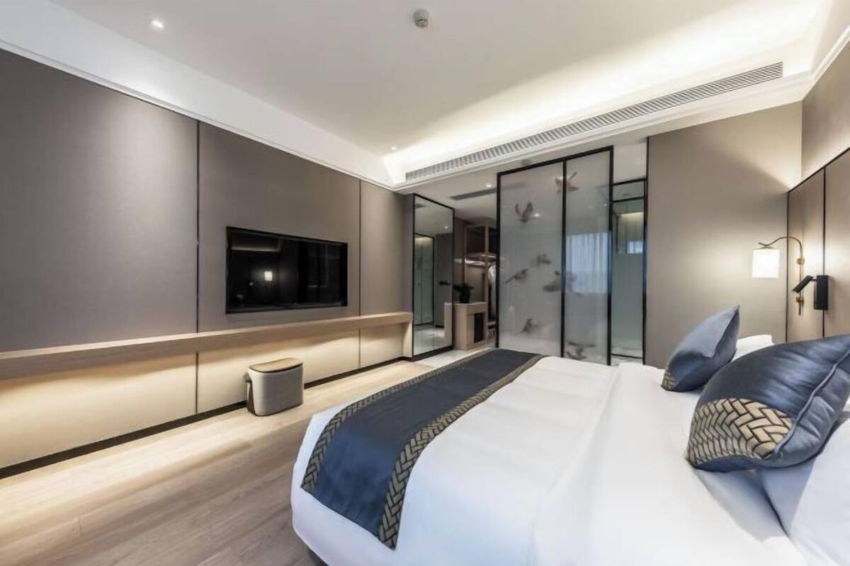 Quarto de hotel moderno com uma cama grande, televisão de tela plana, espelho e uma pequena lixeira.