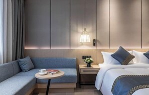 Habitación de hotel con sofá, una pequeña mesa redonda con una taza y un libro, una mesita de noche con un jarrón, y una cama con almohadas.