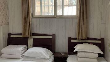 A room with two single beds, each with a wooden headboard and white bedding. There is a window with curtains, and the walls are paneled.