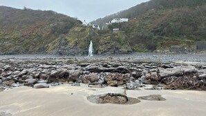 A rocky beach with a waterfall in the background and houses on a hillside. - The Boathouse (Bucks Mills)