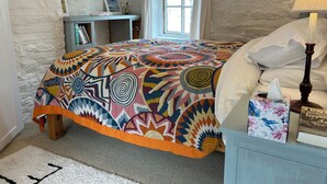 A bedroom with a colorful bedspread, a bedside table with a lamp, a window with curtains, and a bookshelf with books. - The Boathouse (Bucks Mills)