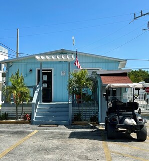 A small, single-story building with a light blue exterior, a flagpole with an American flag, a small porch with steps, and a golf cart parked in front. - Key Largo, Charming 2-bedroom RV in Calusa Campground (Key Largo)