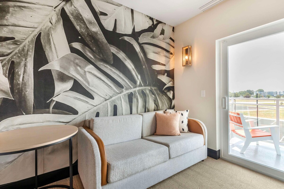 A modern living room with a grey sofa, a round table, and a large abstract leaf mural on the wall.