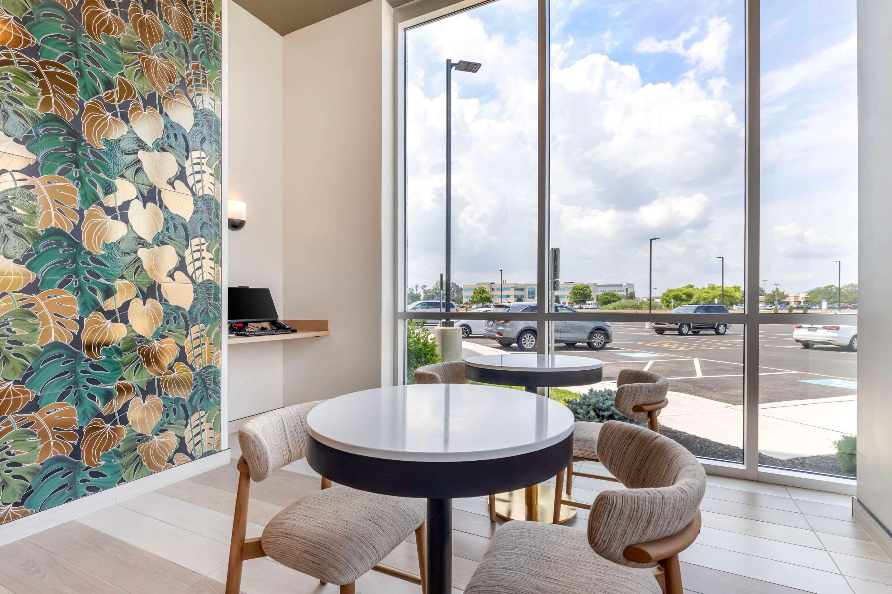 A modern interior with round tables and chairs, a large window with a view of a parking lot and buildings, and a wall with a leafy pattern.