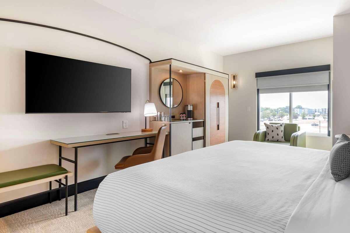 Ein Hotelzimmer mit einem großen Bett, einem Schreibtisch mit Stuhl, einem Fernseher, einem Fenster mit Aussicht und einer kleinen Küchenzeile.