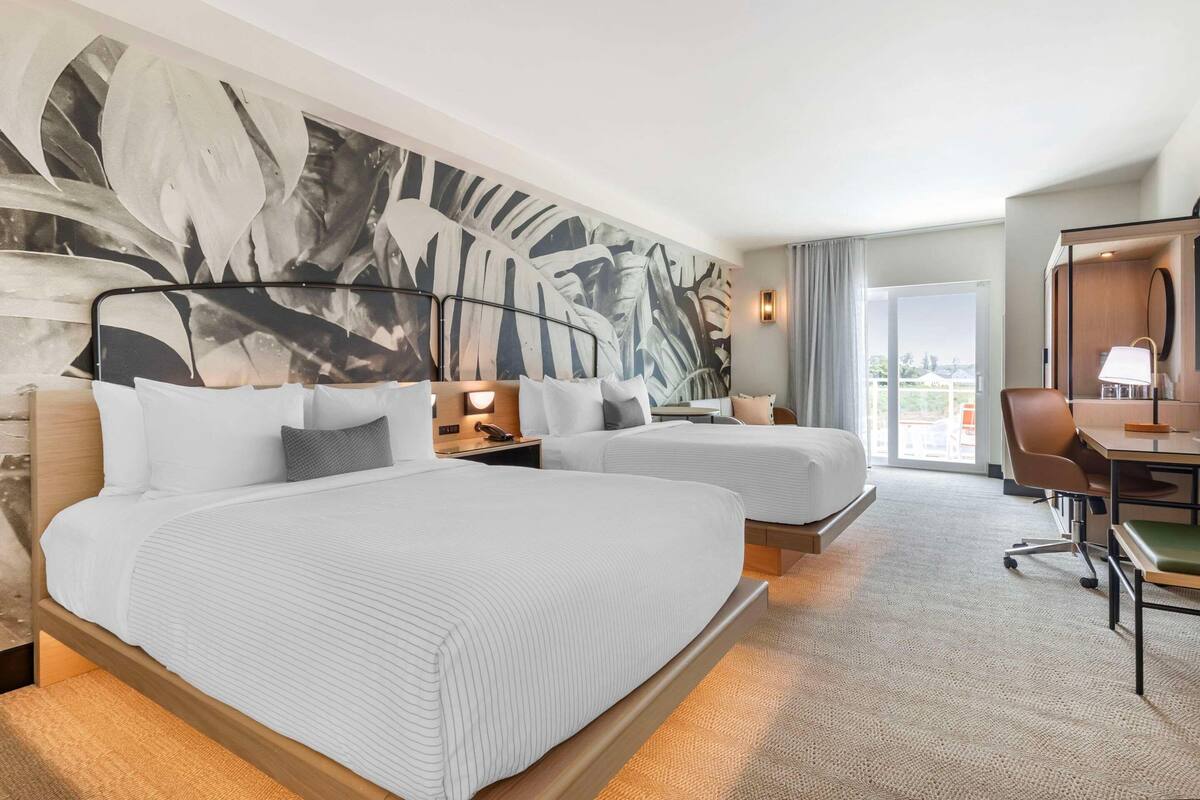 A hotel room with two beds, a desk, a chair, and a large window.
