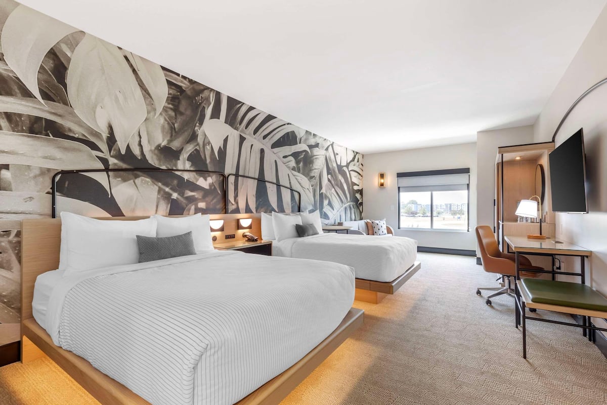 A hotel room with two beds, a desk, a chair, a large abstract wall mural, and a TV.