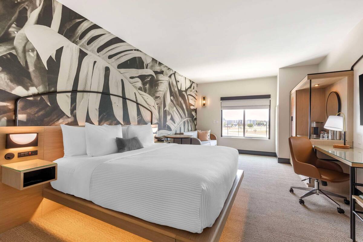 A hotel room with a large bed, a desk, a chair, a nightstand, and a wall-mounted lamp.